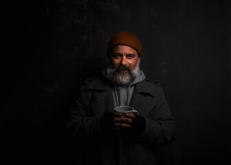 Bearded beggar man smiling, happy even living on the street. Cheerful homeless man with cup of hot tea isolated on dark background