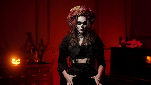 woman in rags witch costume for halloween with calavera catrina skull makeup, horns and dried flowers on her head in a dark hot red room screaming raising her hands and looking at the camera
