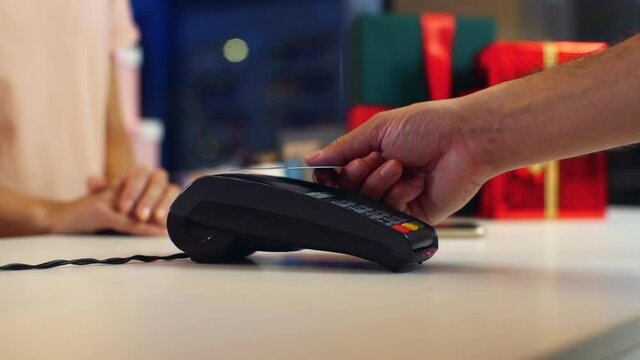 Close-up hands of unrecognizable male customer paying with NFC technology by credit card contactless on POS terminal for Christmas present at holiday store, selective focus, blurred background.