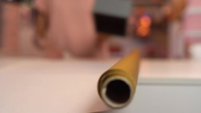 Close up cropped shot of unrecognizable young woman rolling out roll of wrapping golden paper to package festive cardboard box with Christmas present, selective focus. Concept of new year and xmas.