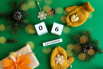 Calendar for December 6 : the name of the month in English, cubes with numbers 0 and 6, yellow mittens, a gift with a yellow bow, spruce branches on a green background, top view, bokeh