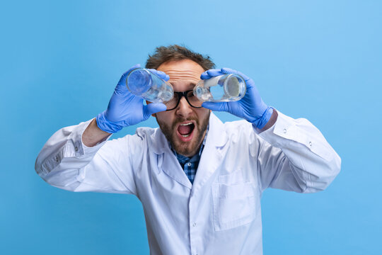 Humorous portrait of young man in image of chemist, doctor wearing white gown and protective gloves isolated on blue background.