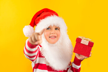 Funny child toddler in a Santa hat with red gift , yellow background. Holidays, Traditions, Christmas concept.