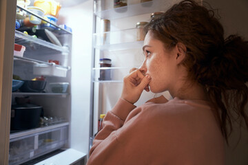 Undecided young caucasian woman checking fridge for some food at night