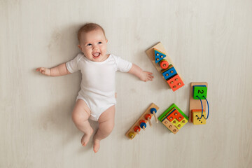cheerful newborn baby in a white bodysuit lies on his back on the floor and plays with educational...