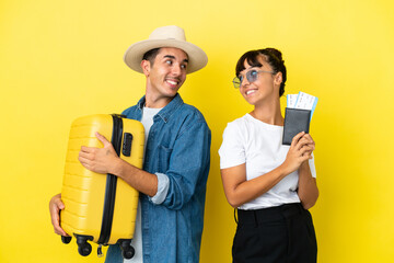 Young traveler friends holding a suitcase and passport isolated on yellow background looking over the shoulder with a smile