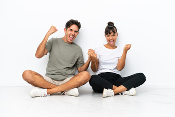 Fototapeta na wymiar Young mixed race couple sitting on the floor isolated on white background celebrating a victory