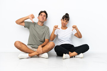 Young mixed race couple sitting on the floor isolated on white background showing thumb down with both hands