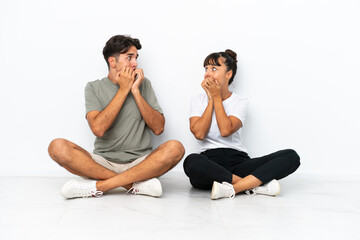 Fototapeta na wymiar Young mixed race couple sitting on the floor isolated on white background is a little bit nervous and scared putting hands to mouth