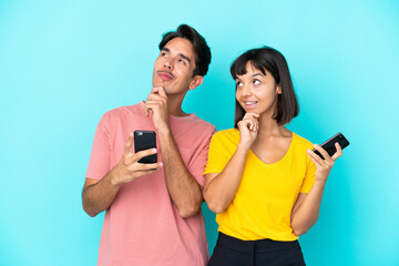 Young mixed race couple holding mobile phone isolated on blue background thinking an idea while looking up