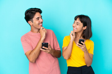 Young mixed race couple holding mobile phone isolated on blue background looking looking at each other