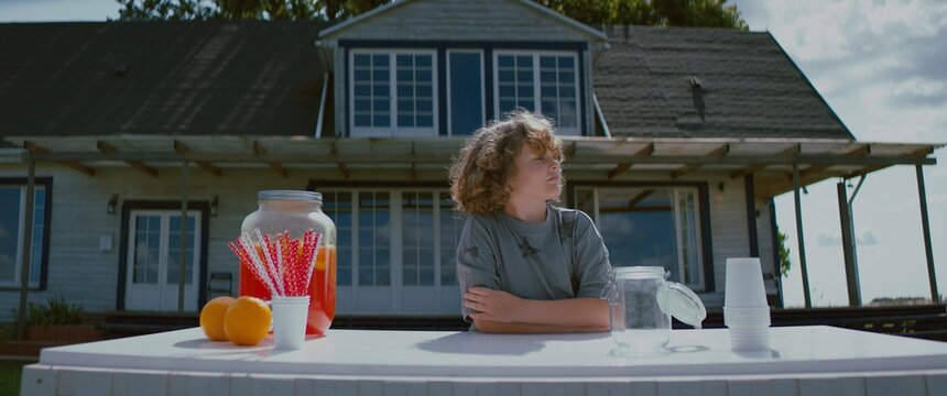 Cute little Caucasian kid boy opens lemonade stand on a lawn in front of his house