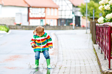 Little toddler boy playing outside on rainy day. Happy positive child running and jumping through rain and puddles. Kid with rain clothes and rubber boots. Children outdoor activity on bad weather day