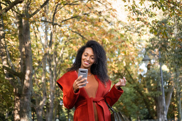 Fototapeta na wymiar Successful elegant black woman receiving good news and texting on her smartphone outside in autumn.