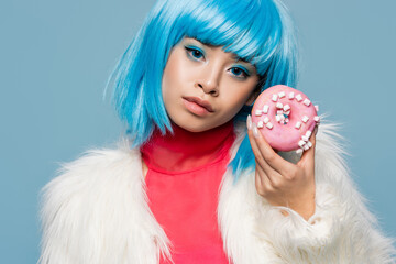 Asian woman in wig and fluffy jacket holding donut and looking at camera isolated on blue