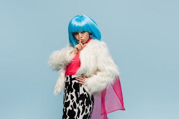 Stylish asian woman in furry jacket showing secret gesture isolated on blue
