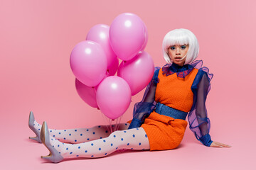 Stylish asian pop art woman looking at camera while sitting near balloons on pink background