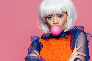 Fashionable asian model blowing bubble gum and looking at camera isolated on pink