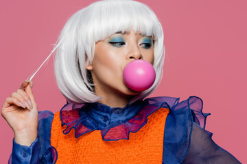 Stylish asian woman touching hair and blowing bubble gum isolated on pink