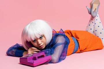 Pretty asian model in white wig lying near retro telephone on pink background