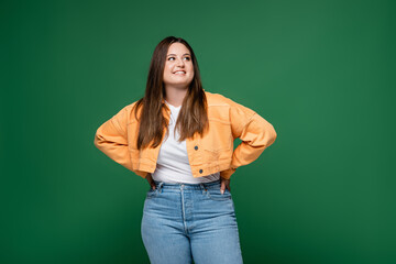 Cheerful plus size woman holding hands on hips isolated on green