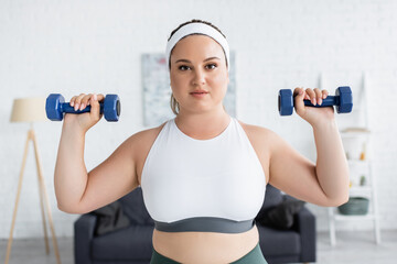 Fototapeta na wymiar Young woman with overweight exercising with dumbbells and looking at camera at home