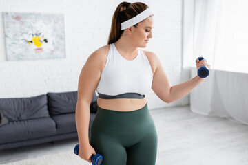 Fototapeta na wymiar Side view of plus size woman training with dumbbells in living room