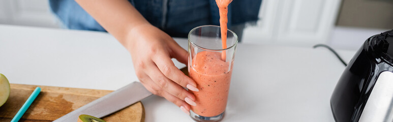 Cropped view of blurred woman pouring smoothie in glass in kitchen, banner