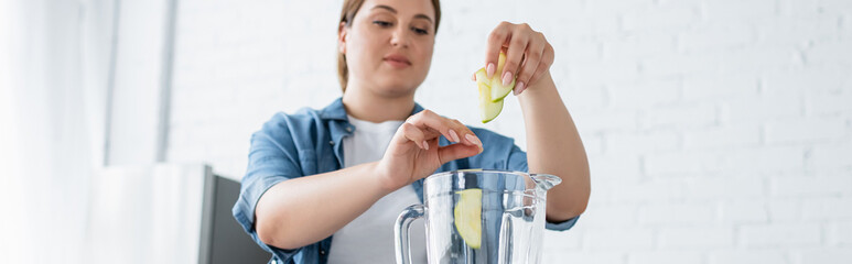 Blurred woman with overweight putting apple slices in blender in kitchen, banner