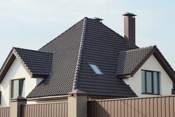 brown tiled roof of a white private house with a window and a chimney against the sky behind a fence wall on the street