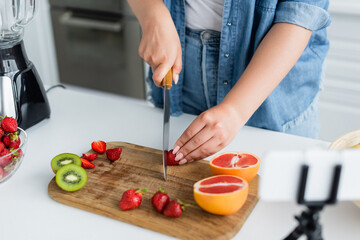 Cropped view of young woman cutting fresh fruits near blender and blurred smartphone in kitchen