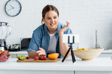 Smiling blogger with overweight looking at smartphone near fresh fruits in kitchen