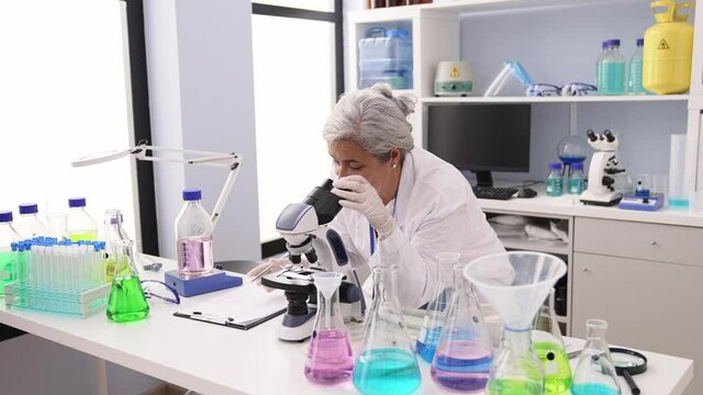 Middle age grey-haired woman wearing scientist uniform using microscope working at laboratory