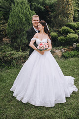 Fototapeta premium A young groom in a black suit and a beautiful brunette bride in a white dress are hugging in the garden, park in nature with green plants. Wedding portrait of newlyweds in love.