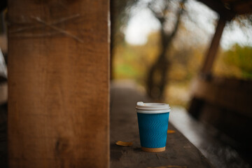 Blue cardboard disposable cup with white plastic lid for coffee. A glass for coffee and hot drinks on a wooden table in a gazebo in an autumn park. Warming drink in the autumn season.