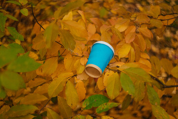 
A blue cardboard disposable cup with a white plastic lid for coffee on a background of yellow leaves. Coffee in the autumn forest. Warming drink in the autumn season.