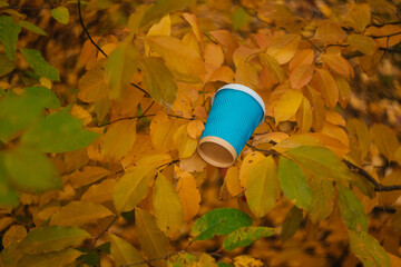 
A blue cardboard disposable cup with a white plastic lid for coffee on a background of yellow leaves. Coffee in the autumn forest. Warming drink in the autumn season.