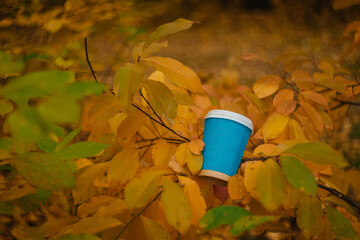 A blue cardboard disposable cup with a white plastic lid for coffee on a background of yellow leaves. Coffee in the autumn forest. Warming drink in the autumn season.