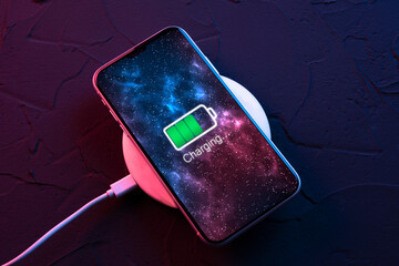 Mobile smart phone on wireless charging device on dark neon red and blue color background. Icon battery and charging progress lighting on screen.smartphones connected to power source.Low battery - obrazy, fototapety, plakaty