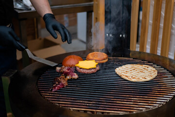 Chef cooking pieces of meat, buns, cutlet with cheese for burgers and pita on grill at summer local...