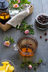 Rose hip tea in glass cup with teapot, dry berries, roses and cut lemon 
