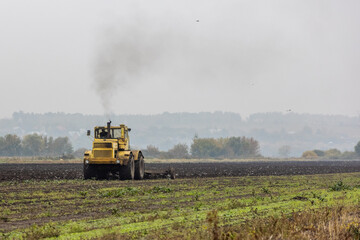 grim yellow tractor plows the field after harvest before winter at cloudy and foggy autumn morning