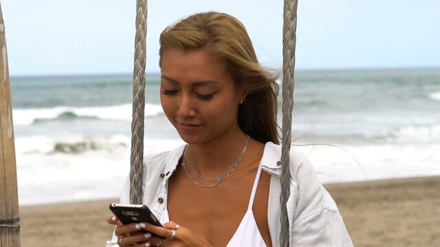 Young, happy woman texting on smartphone sitting on swing at beach