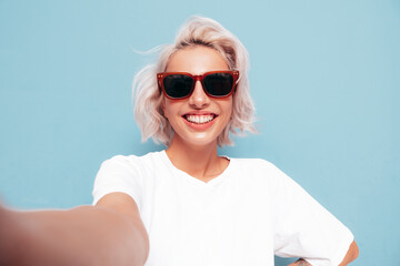 Young beautiful smiling female in trendy summer clothes. Sexy carefree woman posing near blue wall in studio. Positive blond model having fun in sunglasses. Cheerful and happy. Taking Pov selfie