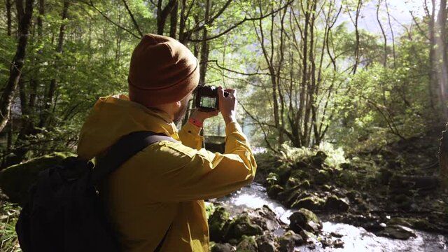 Bearded young adult male tourist in raincoat with camera takes pictures of natural mountain river. Traveler photographer man on vacation trip in highlands among wood tree forest. High land waters.