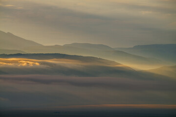 Scenic dawn mountain landscape with golden low clouds in valley among mountains silhouettes under cloudy sky. Vivid sunset or sunrise scenery with low clouds in mountain valley in illuminating color.