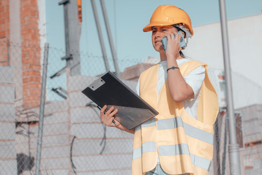 Muchacha de arquitectura estableciendo una comunicaci&oacute;n llamando por tel&eacute;fono m&oacute;vil en la construcci&oacute;n del edificio de obra con sus maquetas y casco de protecci&oacute;n elaborando un plan de estructura