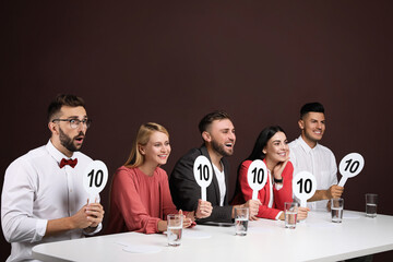 Panel of judges holding signs with highest score at table on brown background - obrazy, fototapety, plakaty