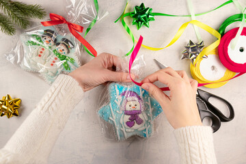 female hands in a white sweater are packing Christmas gifts with gingerbread. gingerbread cookies with snowmen, scissors, wrapping paper, colorful ribbons on the table.