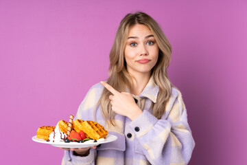 Teenager Russian girl holding waffles isolated on purple background pointing to the laterals having doubts